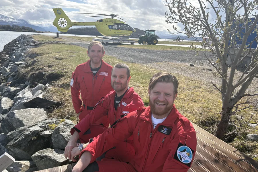 Tre menn i røde uniformer og et gult helikopter i bakgrunnen